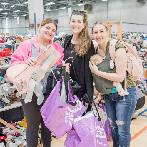 Three generations shop together at their local JBF sale. Mom carries baby in a baby carrier while grandmom stands behind them.