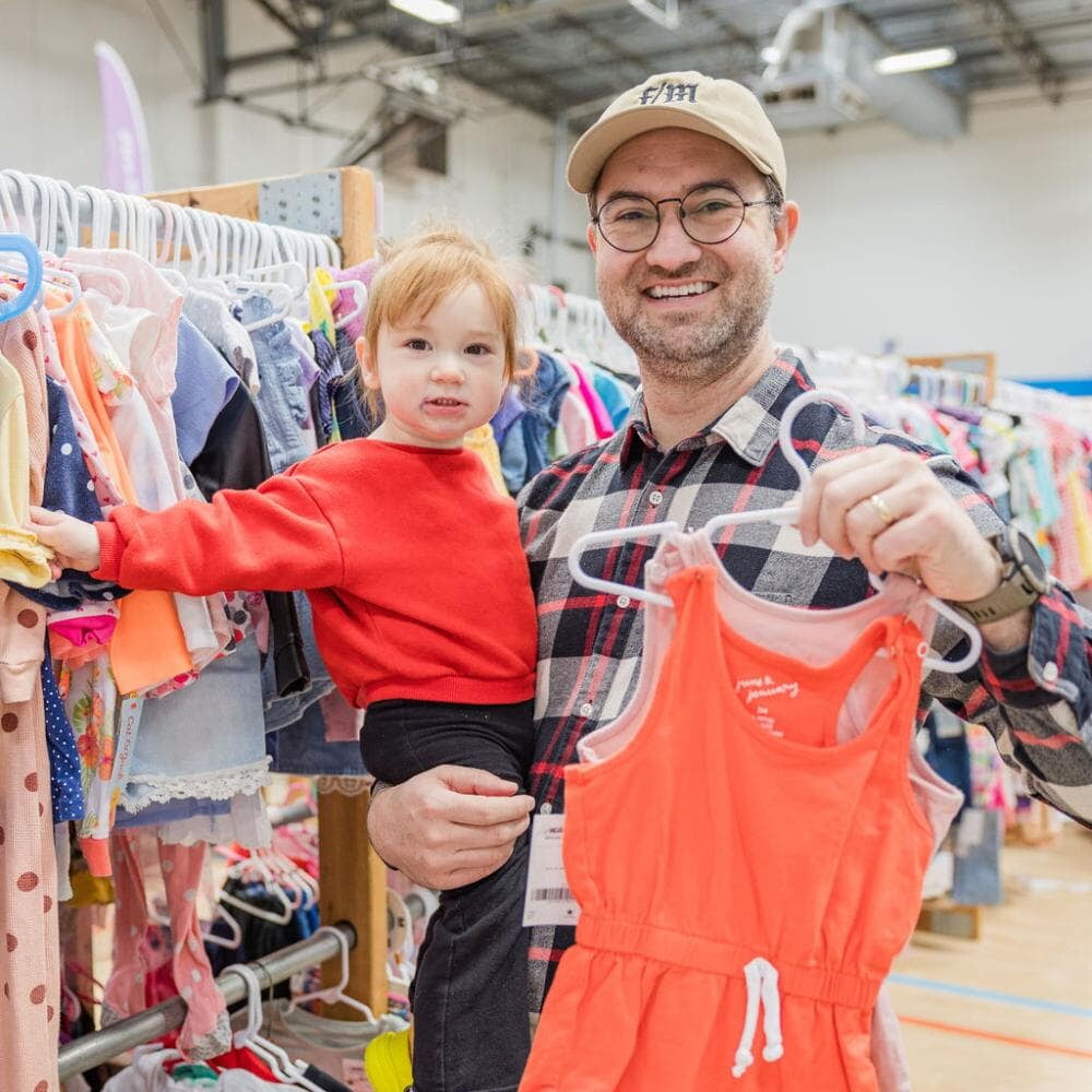 A father smiles and holds his daughter in one hand and a hanger of clothing in the other.