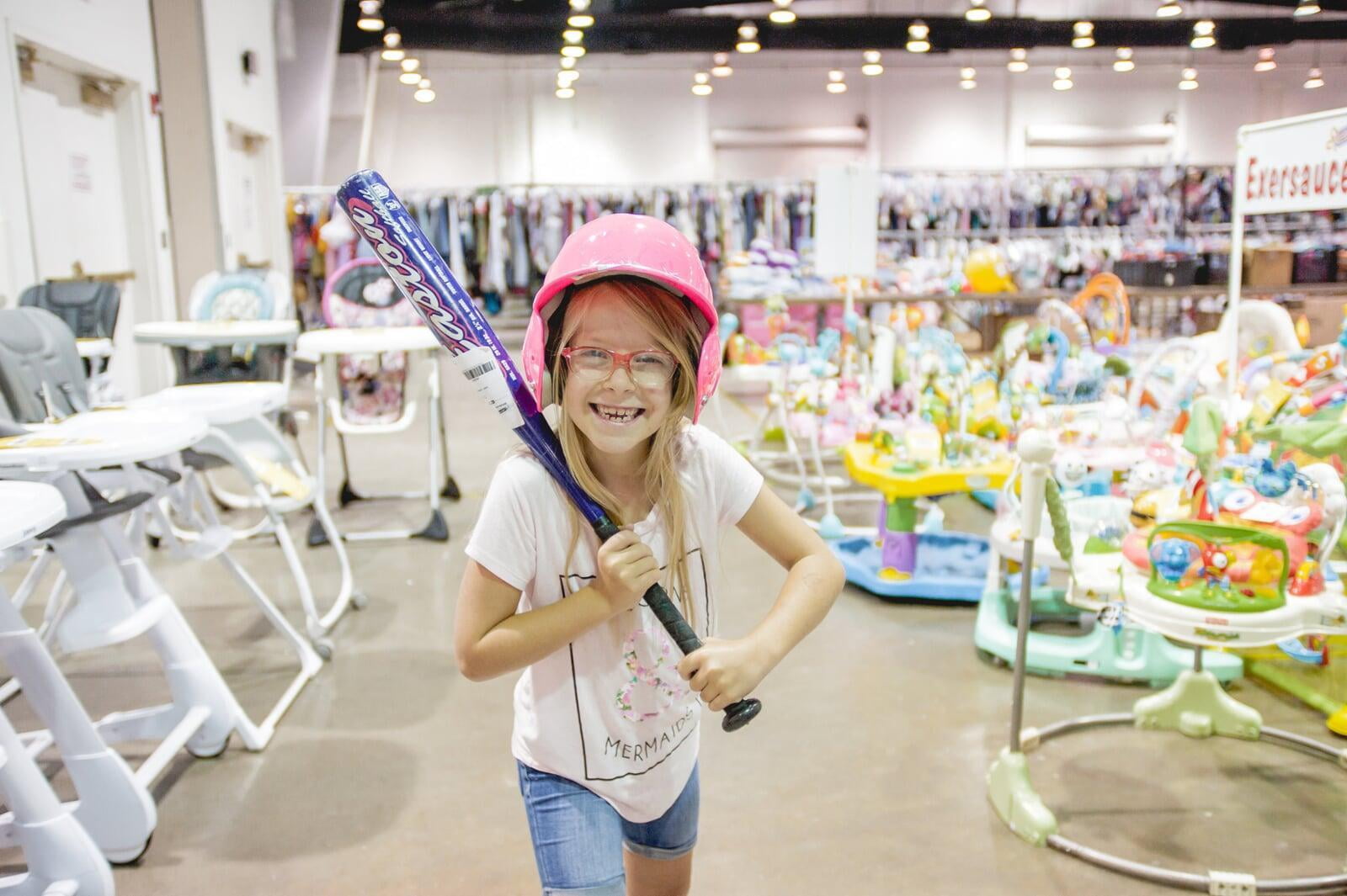 A young girl smiles while holding a baseball bat. She is also wearing a pink baseball hard hat.