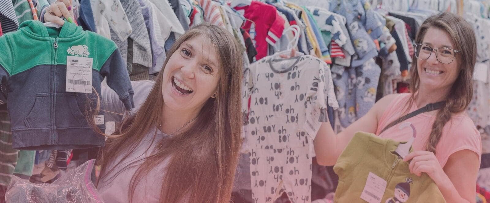 Two ladies smile as the hold up hangers with clothing on them. They are both standing next to a rack full of clothing for sale.